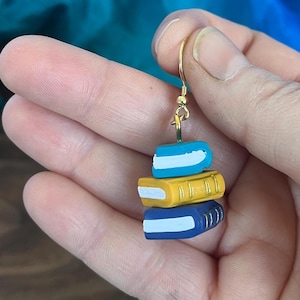 May include: A dangle earring featuring a stack of three miniature books in bright blue, yellow, and dark blue. Each book has a white page edge and gold-tone spine detailing. The stack hangs from a gold-tone metal hook for pierced ears.
