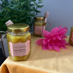 May include: A jar of Busy B's Homemade Honey Lemon Scrub with a gold lid and pink label. A pink flower and a wooden honey dipper are also visible. The jar is on a yellow surface.