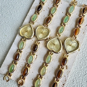May include: Four gold-toned quartz watches with heart-shaped faces and linked bands. The bands feature alternating oval-shaped green and brown stones. The watch faces have gold hands and hour markers. The watches are displayed on a light-colored surface.