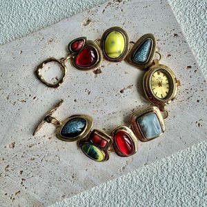 May include: A vintage gold-toned bracelet featuring a variety of colorful, gem-like stones in red, yellow, blue, and green. The bracelet includes a small, round watch with a gold face. The bracelet is approximately 7 inches in length.