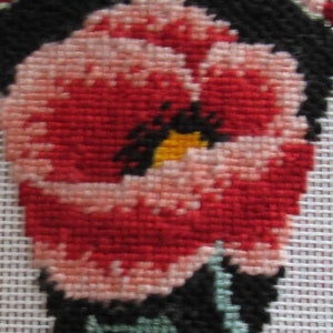 May include: Needlepoint design of a stylised red and pink flower with black accents and green leaves. The flower has a yellow centre and is set against a white background. The top edge is finished with a beaded trim.