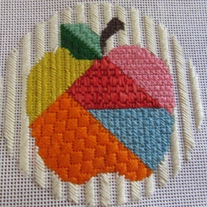 May include: A colourful needlepoint design of an apple with a green leaf and brown stem. The apple is made up of six different coloured sections: yellow, orange, red, pink, blue and green.