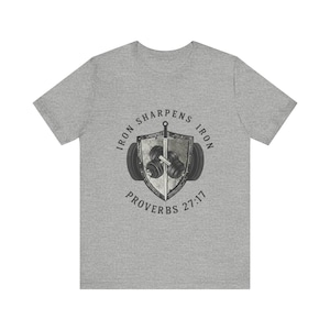 Puede incluir: Camiseta gris con un diseño de escudo, una espada y pesas. El texto "IRON SHARPENS IRON" se arquea sobre el escudo, con "PROVERBIOS 27:17" debajo. El diseño es en tonos grises y negros.