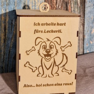 Könnte beinhalten: Eine hellfarbene Holzkiste mit einem pfotenförmigen Deckelgriff. Die Kiste zeigt einen lasergravierten Cartoon-Hund, der von Knochen und deutschem Text umgeben ist. Der Text lautet: "Ich arbeite hart für das Leckerli. Also... hol schon eins raus!"