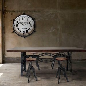 May include: An industrial-style dining table with a dark wood top and metal legs, paired with three adjustable stools. A large, round clock with the text "Grand Central Station New York" hangs on the wall.