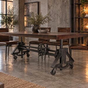 May include: A rectangular wooden dining table with a dark brown finish, supported by a heavy-duty black metal industrial base featuring visible gears, a hand crank, and caster wheels, accompanied by four brown leather chairs with metal frames.