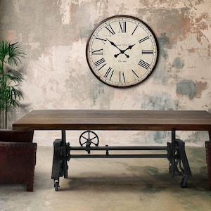 May include: A rustic interior scene featuring a large wooden table with a metal base, flanked by two leather armchairs. A large round clock with Roman numerals hangs on the wall. A green plant adds a touch of nature.