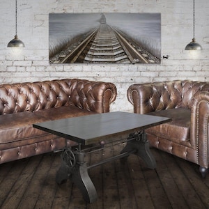 May include: A living room setting featuring two brown leather Chesterfield sofas and a matching coffee table with a metal base. The room has a rustic feel with a painting of train tracks and hanging pendant lights.