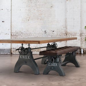 May include: A rustic wooden dining table with a metal base featuring adjustable crank wheels. The table is paired with a matching bench with a brown leather seat. The table and bench are labeled "KNOX".