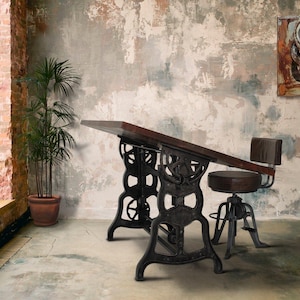 May include: An industrial-style drafting table with a dark wood top and ornate cast iron base. A matching adjustable stool with a brown leather seat sits beside it. A potted plant and artwork complete the room.