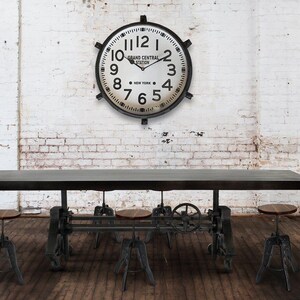 May include: Industrial-style dining table with three matching stools. A large clock with the words "Grand Central Station New York" hangs on a white brick wall. Two potted plants flank the table, creating a balanced composition.