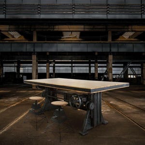 May include: Industrial-style dining table with a light-colored wooden top and a dark metal base. The table features a hand crank mechanism and is paired with two small, round stools. The setting is a large, open industrial space.