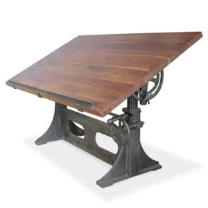May include: A vintage-style drafting table with a solid wood top and a cast iron base. The tabletop is angled and adjustable. The base features a hand crank and gear system for height adjustment. The wood has a warm brown tone.