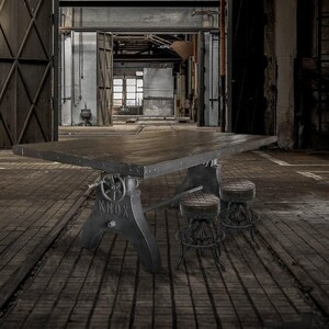 May include: A large, industrial-style table with a dark, weathered finish. The table has a rectangular top and is supported by a metal base with adjustable features. Two small stools are positioned in front of the table. The setting is an old warehouse.