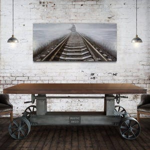May include: A rustic industrial dining table with a wooden top and metal base. The table is on wheels and has a metal lever to adjust the height. Two gray upholstered chairs are placed on either side of the table. A framed print of a railroad track in a foggy landscape hangs on the wall behind the table.
