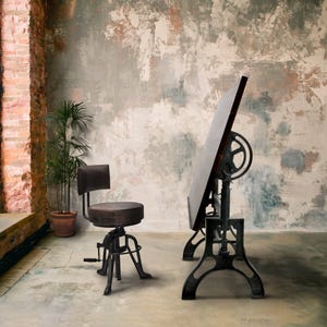 May include: An industrial-style drafting table with a large, angled wooden surface and a cast-iron base. A matching adjustable stool with a round seat and backrest sits nearby. The scene is set against a textured, aged wall and a brick column.