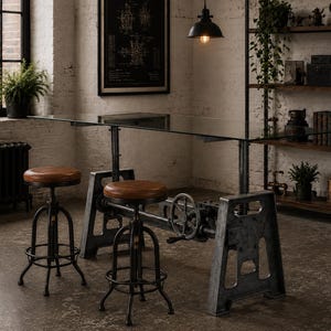 May include: A rectangular glass-top table with a heavy-duty iron base featuring a mechanical hand crank for height adjustment, accompanied by two round stools with brown leather seats and black metal frames with circular footrests.