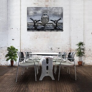 May include: A silver metal table with a square top and a unique, industrial-style base. The table is surrounded by six silver chairs with gray cushions. A black and white painting of an airplane hangs on the wall behind the table.