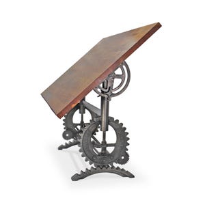 May include: An industrial-style drafting table with a wooden top and a metal base. The base features large gears and a crank mechanism for adjusting the table's height and angle. The table's surface is a warm brown color, and the metal has a dark, aged finish.