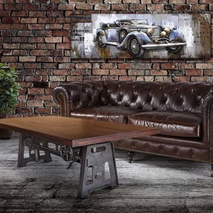 May include: A living room scene featuring a brown leather Chesterfield sofa, a wooden coffee table with metal legs, and a vintage car artwork on a brick wall. Potted plants add a touch of greenery.