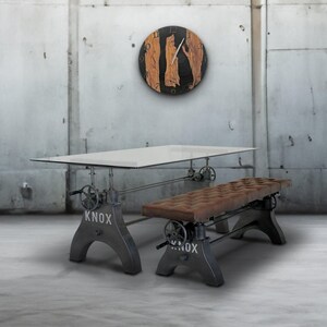 May include: Industrial-style dining table with a glass top and metal base, paired with a matching bench. The table and bench have a distressed finish and the word "KNOX" is printed on the base. A round clock with wood and black accents hangs on the wall.