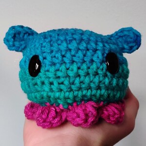 Puede incluir: Una criatura de ganchillo hecha a mano con un cuerpo redondeado, dos orejas pequeñas y grandes ojos de botón negros. La parte superior es azul, que pasa a verde azulado, con una base festoneada magenta. Este juguete de peluche suave es una creación única y caprichosa.