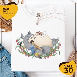 a white t-shirt with a colorful illustration of a cat and various other characters, including a blue cat, surrounded by flowers and leaves. The t-shirt is displayed on a white background, and there are yellow and black sale signs visible in the image.