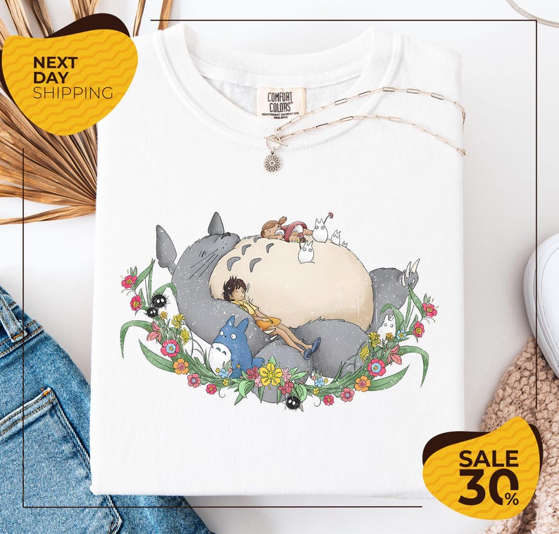 a white t-shirt with a colorful illustration of a cat and various other characters, including a blue cat, surrounded by flowers and leaves. The t-shirt is displayed on a white background, and there are yellow and black sale signs visible in the image.
