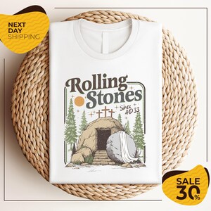 a white t-shirt with a graphic design depicting a rock with a cave entrance, surrounded by trees and a cross. The text on the t-shirt reads "Rolling Stones Since 4D33". The t-shirt is displayed on a woven placemat, and there are two yellow sale signs in the image, one on the left and one on the right, indicating a sale or discount.