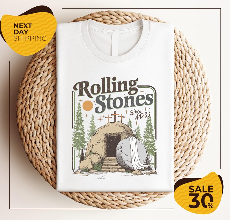 a white t-shirt with a graphic design depicting a rock with a cave entrance, surrounded by trees and a cross. The text on the t-shirt reads "Rolling Stones Since 4D33". The t-shirt is displayed on a woven placemat, and there are two yellow sale signs in the image, one on the left and one on the right, indicating a sale or discount.