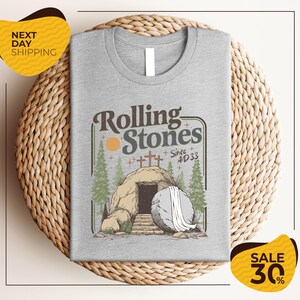 a gray t-shirt with a graphic design depicting a rock with an entrance to an underground passage, surrounded by trees and a cross. The text on the t-shirt reads "Rolling Stones Since 4D33" and there are two yellow sale signs in the image, one on the left and one on the right, indicating a 30% discount on the t-shirt.