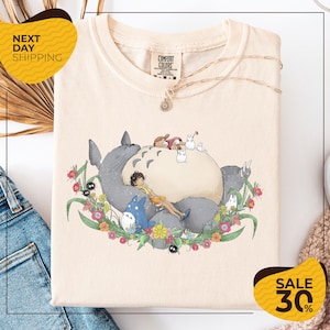a white t-shirt with a colorful graphic design depicting a character surrounded by various animals and flowers. The t-shirt is displayed on a white background, and there are yellow and black sale signs indicating a 30% discount.
