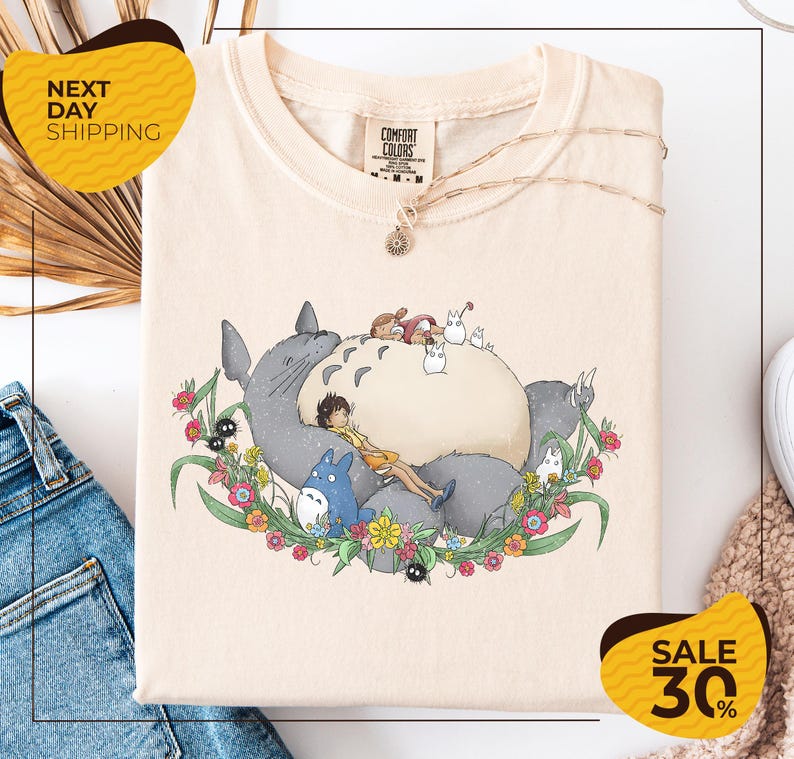 a white t-shirt with a colorful graphic design depicting a character surrounded by various animals and flowers. The t-shirt is displayed on a white background, and there are yellow and black sale signs indicating a 30% discount.