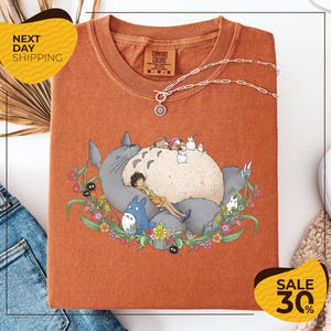 a red t-shirt with a cartoon illustration of a cat and various other characters, such as a blue bird and a gray cat, surrounded by flowers and leaves. The t-shirt is displayed on a white background, and there are yellow and black sale signs indicating a 30% discount.