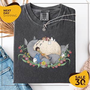 a gray t-shirt with a graphic design depicting a cat surrounded by various animals and flowers. The t-shirt is displayed on a white background, and there are some jeans and a pair of sunglasses visible in the image.