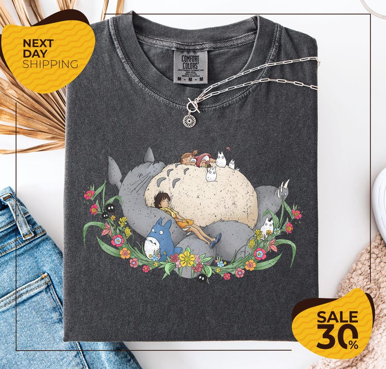 a gray t-shirt with a graphic design depicting a cat surrounded by various animals and flowers. The t-shirt is displayed on a white background, and there are some jeans and a pair of sunglasses visible in the image.