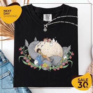 a black t-shirt with a colorful graphic design depicting a cat surrounded by various animals and flowers. The t-shirt is displayed on a white background, and there are some jeans and a necklace visible in the background.