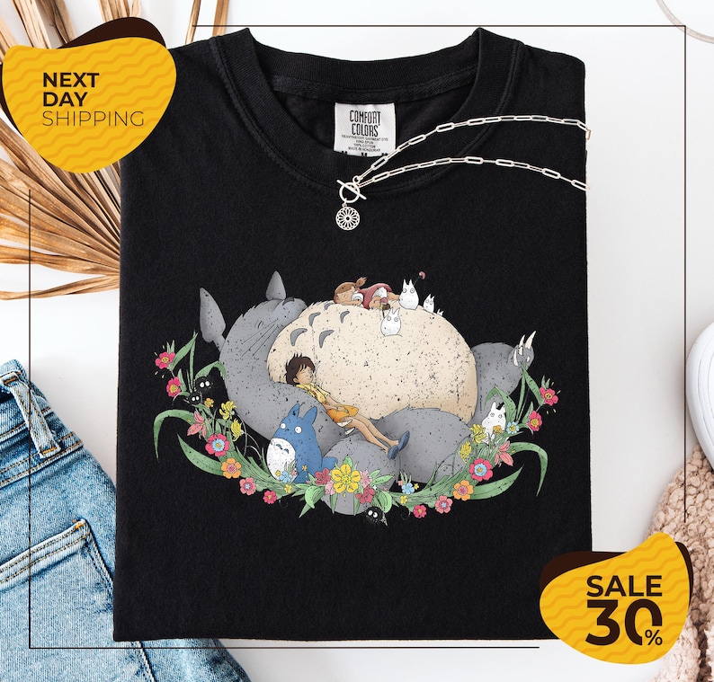a black t-shirt with a colorful graphic design depicting a cat surrounded by various animals and flowers. The t-shirt is displayed on a white background, and there are some jeans and a necklace visible in the background.