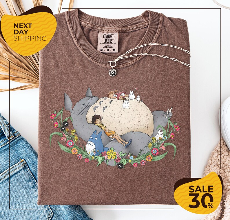 a brown t-shirt with a graphic design depicting a cat surrounded by various cartoon-style animals and flowers. The t-shirt is displayed on a white background, and there are yellow and black sale signs indicating a 30% discount.