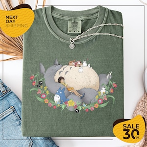 a green t-shirt with a colorful illustration of a cat surrounded by various animals and flowers, along with a necklace. The t-shirt is displayed on a white background, and there are some jeans and a sweater visible in the background.