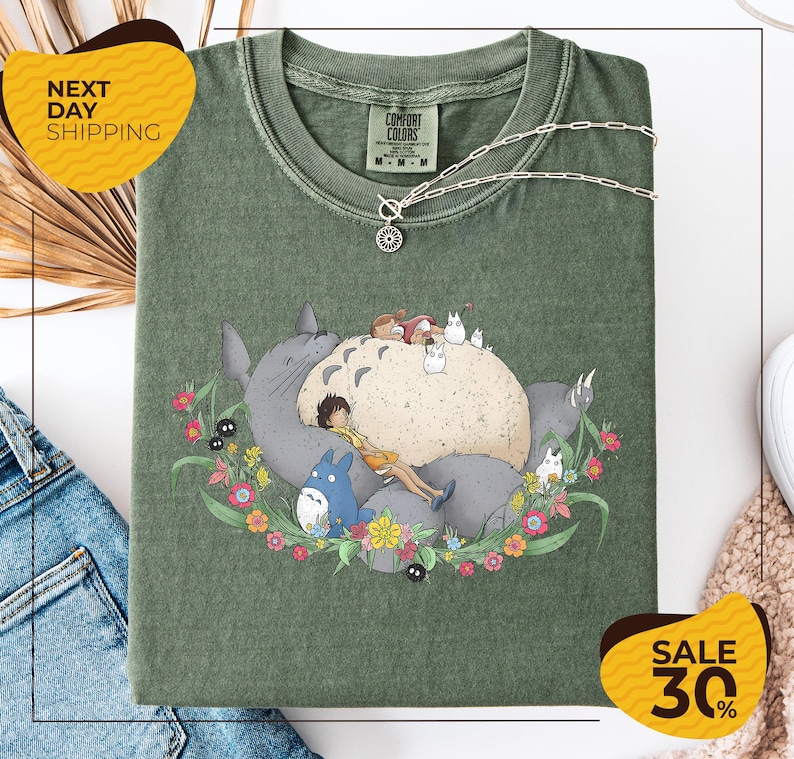 a green t-shirt with a colorful illustration of a cat surrounded by various animals and flowers, along with a necklace. The t-shirt is displayed on a white background, and there are some jeans and a sweater visible in the background.