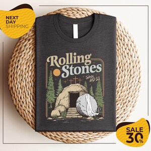 a gray t-shirt with a graphic design depicting a rock with a cave entrance, surrounded by trees and a cross. The text on the t-shirt reads "Rolling Stones Since #33". The t-shirt is displayed on a woven placemat, and there are two yellow price tags in the image, one for "SALE 30%" and another for "NEXT DAY SHIPPING".