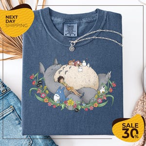 a blue t-shirt with a colorful graphic design depicting a cat surrounded by various animals and flowers. The t-shirt is displayed on a white background, and there are yellow and black sale signs indicating a 30% discount.