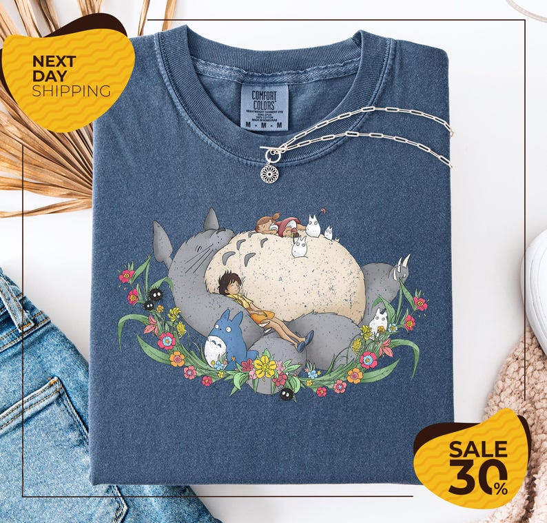 a blue t-shirt with a colorful graphic design depicting a cat surrounded by various animals and flowers. The t-shirt is displayed on a white background, and there are yellow and black sale signs indicating a 30% discount.