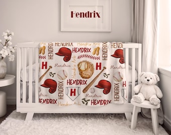 Op de afbeelding: Een witte babydeken met een honkbalthema, met rode honkbalhelmen, knuppels, handschoenen en honkballen. De naam "Hendrix" is rood gedrukt in het hele ontwerp. Een ingelijste print boven de wieg toont ook de naam "Hendrix".