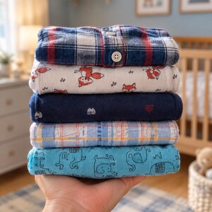Op de afbeelding: Een stapel van vijf opgevouwen babyoverhemden. Het bovenste overhemd is marineblauw, rood en wit geruit met knopen. Het tweede is wit met vossenillustraties. Het derde is effen marineblauw. Het vierde is lichtblauw geruit. Het onderste is lichtblauw met olifantenprints.