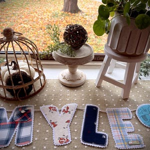 Op de afbeelding: Een stoffen banner met de naam MYLES, waarbij elke letter is gemaakt van verschillende textielsoorten met patronen, waaronder ruitjes, effen marineblauw en een lichtblauwe olifantenprint, uitgestald naast een draadkooi en plant.