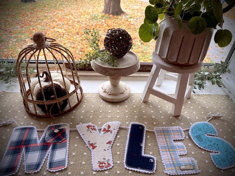 Op de afbeelding: Een stoffen banner met de naam MYLES, waarbij elke letter is gemaakt van verschillende textielsoorten met patronen, waaronder ruitjes, effen marineblauw en een lichtblauwe olifantenprint, uitgestald naast een draadkooi en plant.