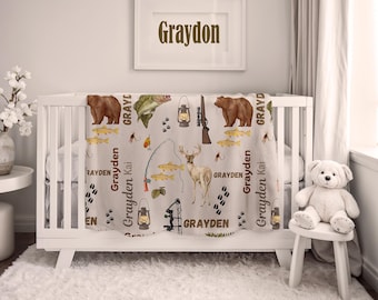 Op de afbeelding: Een babydeken met een jachtthema, met illustraties van een beer, hert, vis en visgerei. De deken is over een witte wieg gedrapeerd. De naam "Grayden" staat op de deken en in een ingelijste foto.