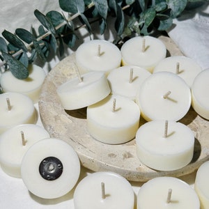May include: A close-up of a group of white tea light candles with wicks on a light brown surface. The candles are arranged in a circle with a few candles overlapping.